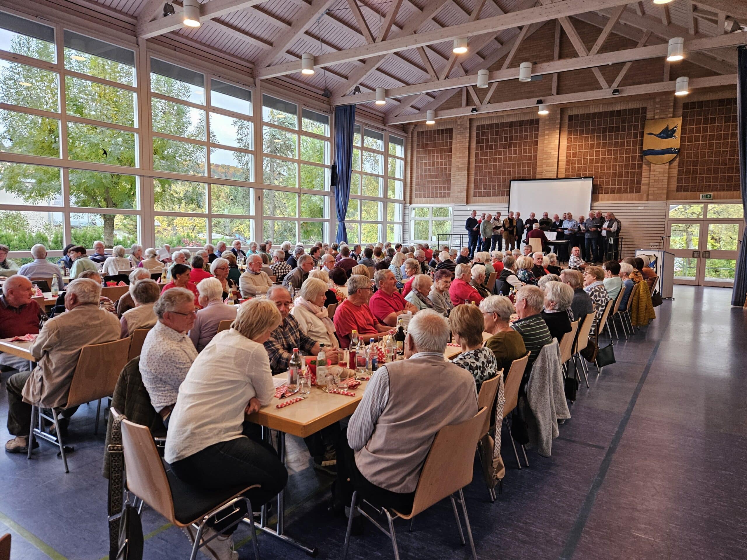 Mehrere Senioren sitzen an den Tischen und eine Gruppe Älterer Herren tragen etwas vor